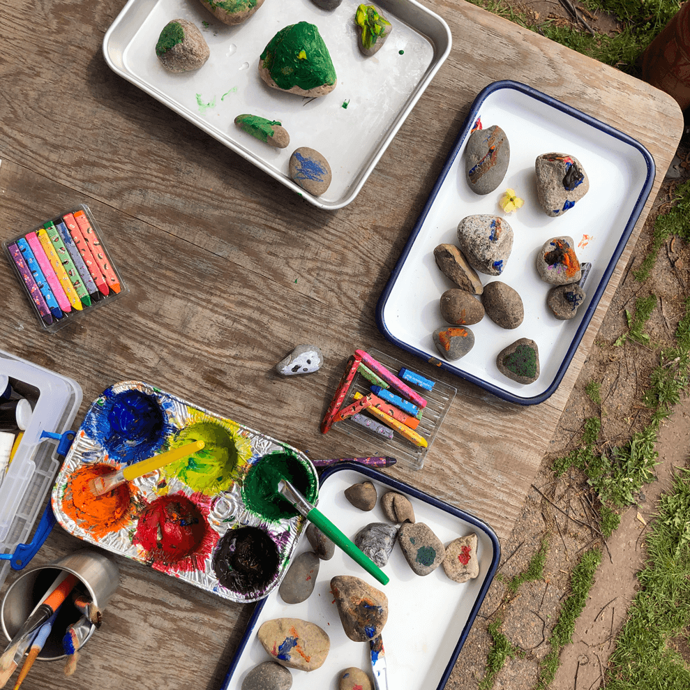 A collection of painting supplies and painted rocks are spread out on a wooden table outdoors. There are trays containing rocks painted in different colors, a palette with various paints and brushes, and a set of oil pastels.