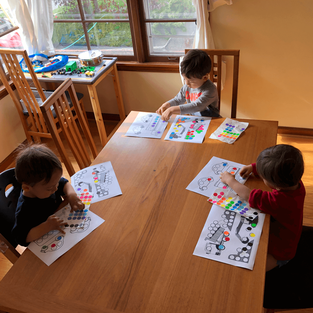Three young children are seated at a wooden table, each working on coloring activities with dot stickers. The table is set in a brightly lit room with large windows in the background.